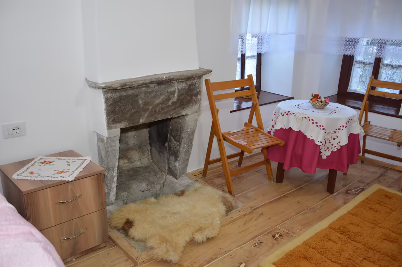 Corner of a room with a stone fireplace, a small table with two wooden chairs and a bedside table