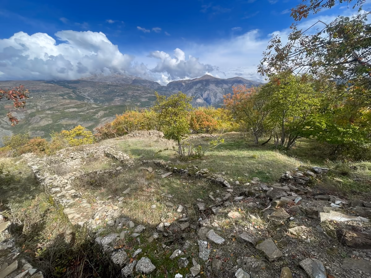 Outlines of old stone walls on a grassy terrace, with trees and cloudy mountains behind