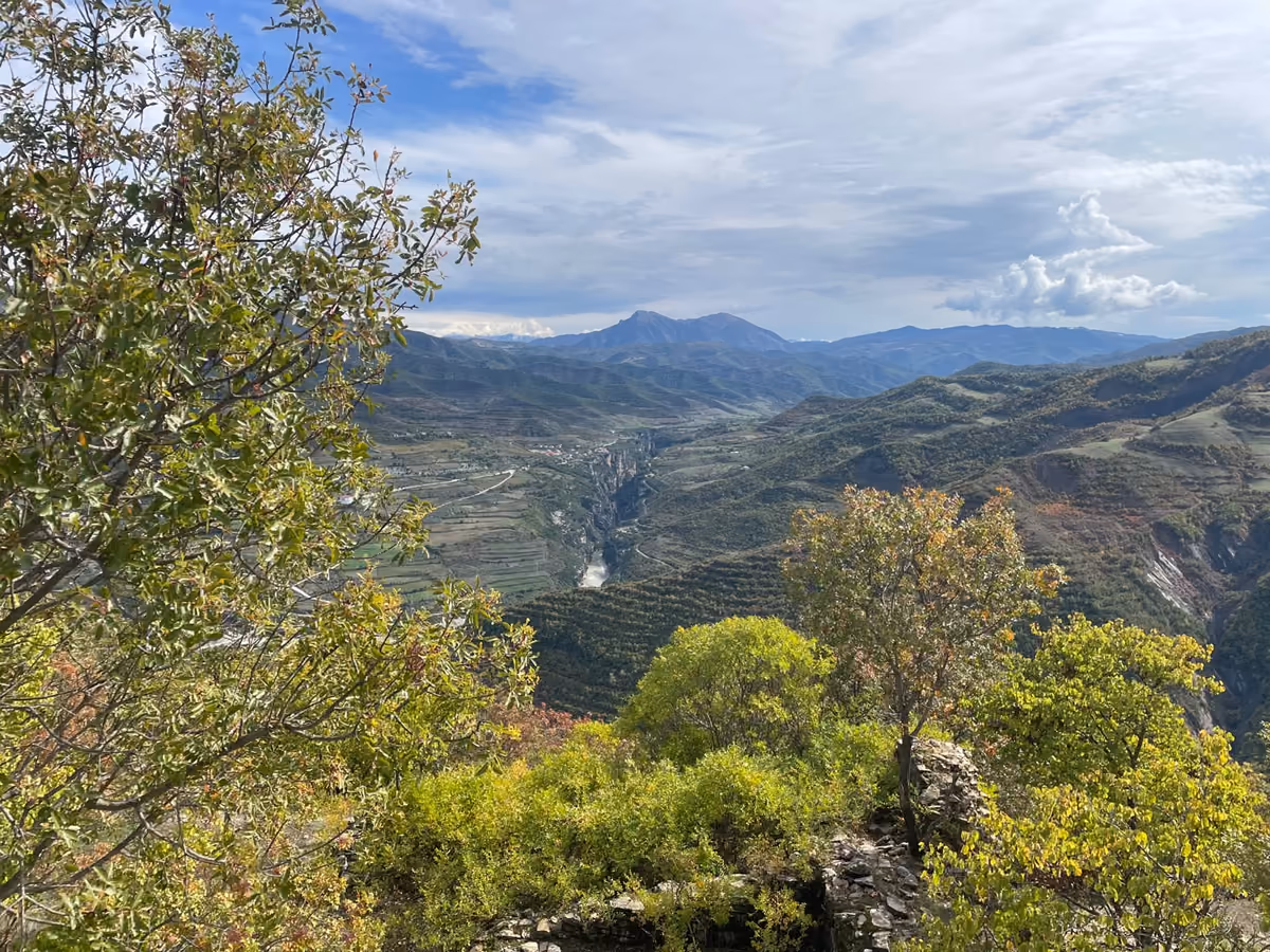 View through trees to a river gorge and a pointed peak, with a corner of the old walls in the foreground