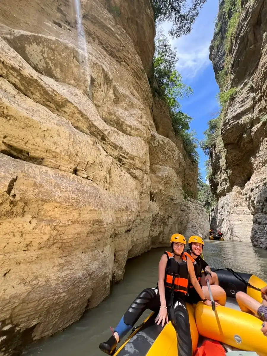Two rafters in yellow helmets below a golden cliff with a trickling waterfall